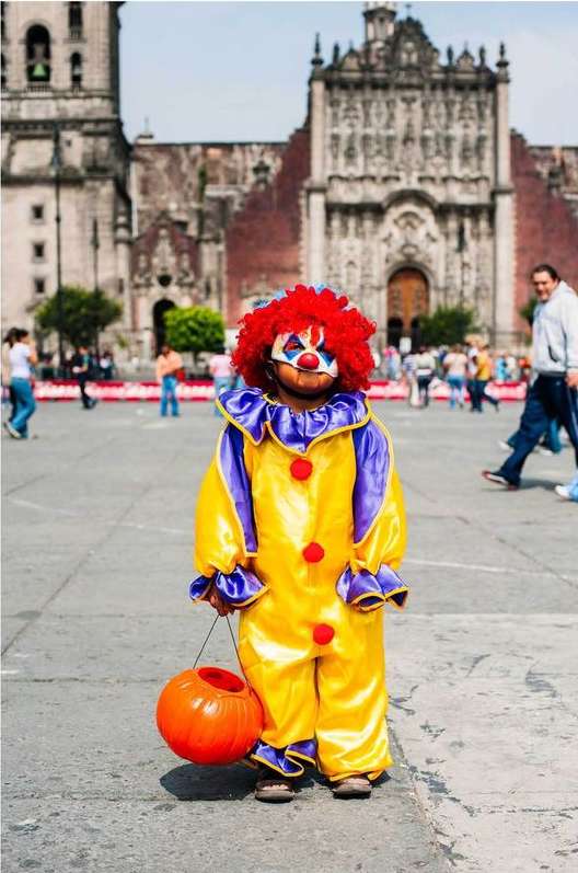 Child in colorful clown costume holds pumpkin bucket in urban square with blurred background featuring people and a large building.