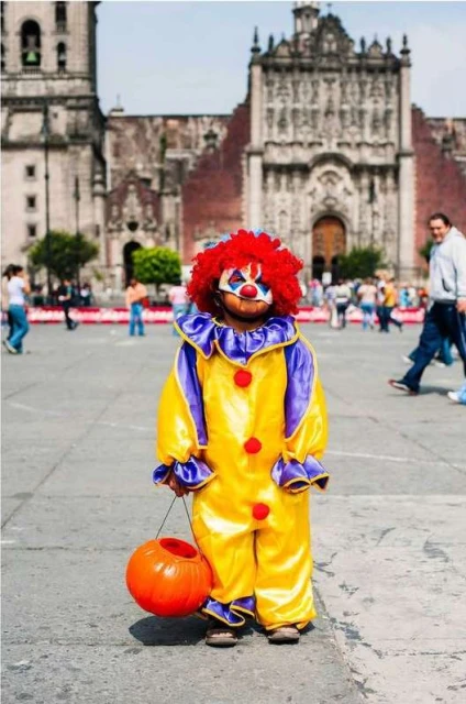 Child in colorful clown costume holds pumpkin bucket in urban square with blurred background featuring people and a large building.