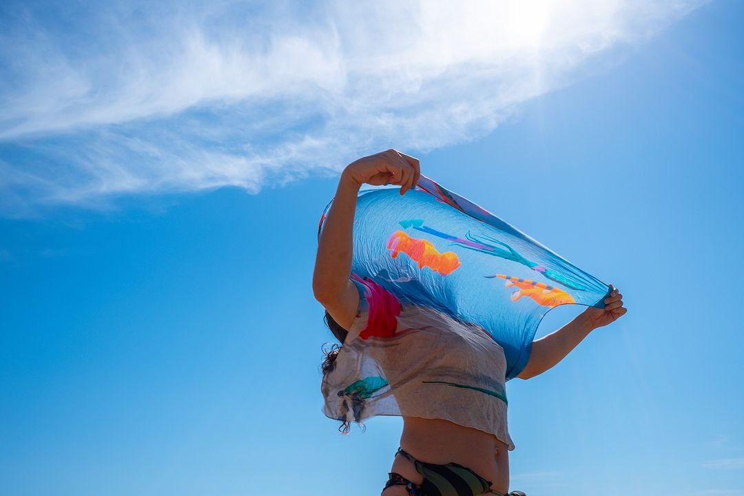 A woman holds a translucent scarf with colorful fish designs up to a sunny, partly cloudy blue sky, creating a vibrant and airy composition.