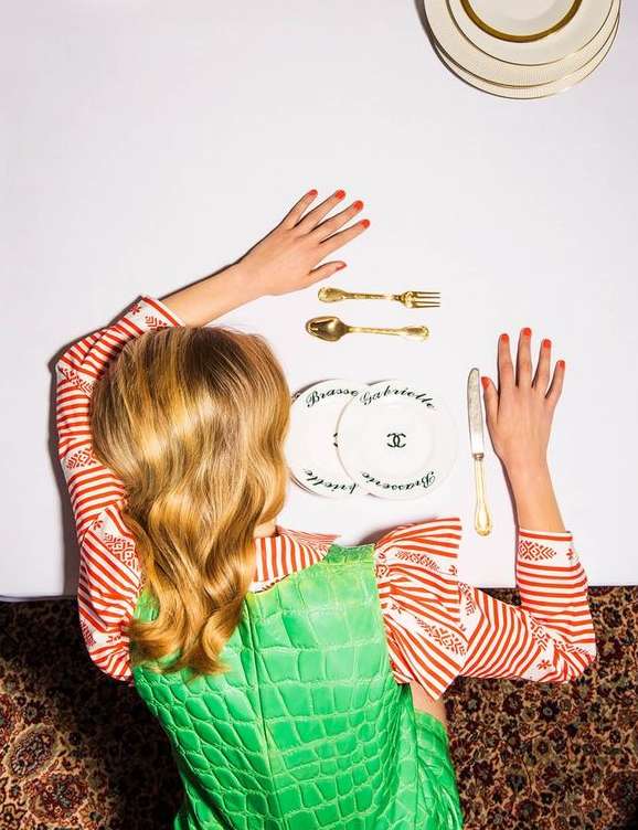 Overhead shot of a fair-skinned woman in a patterned top and green vest lying on a floral patterned surface near dining flatware and plates.
