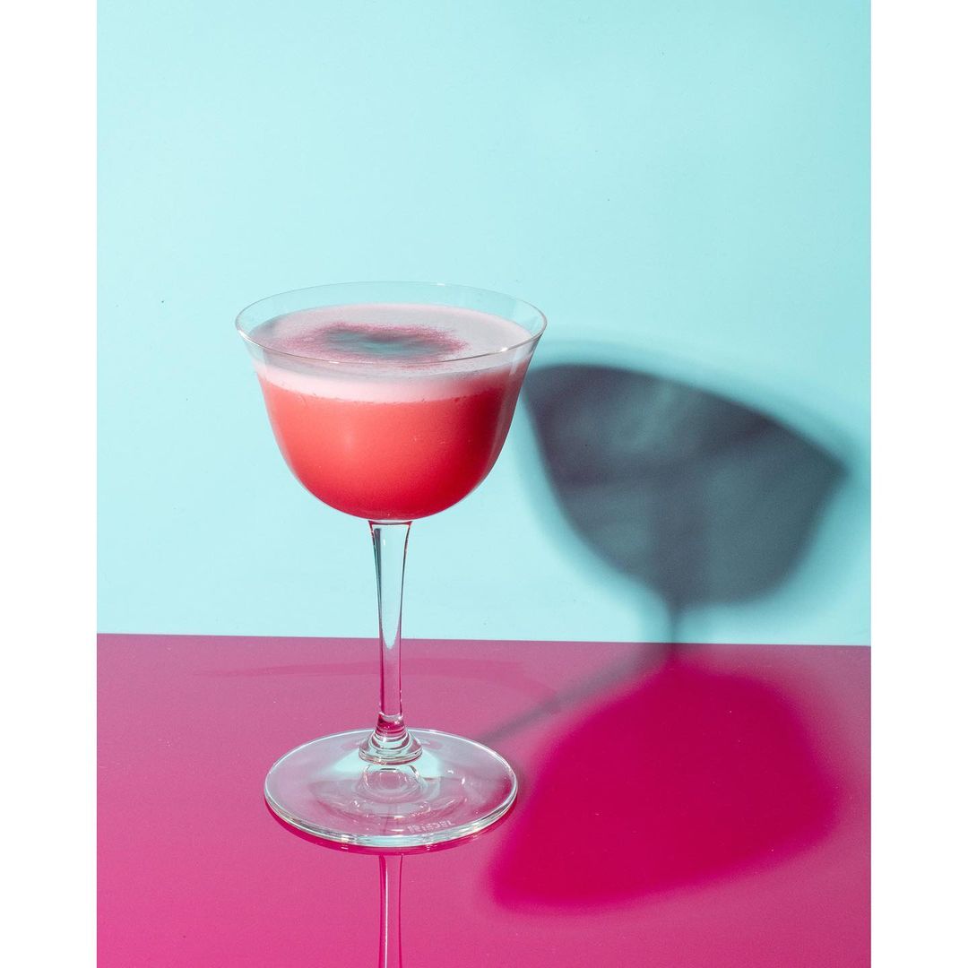 Elegant stemmed glass filled with a vibrant pink cocktail with a foamy white top, set against a contrasting blue and pink backdrop.