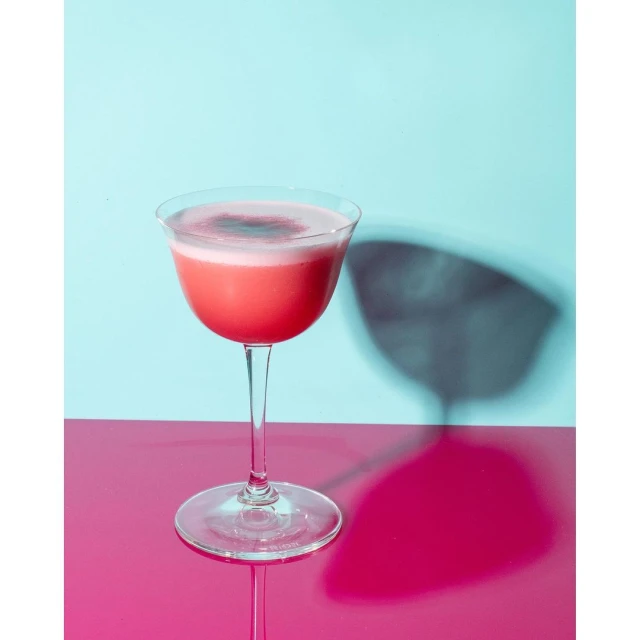 Elegant stemmed glass filled with a vibrant pink cocktail with a foamy white top, set against a contrasting blue and pink backdrop.