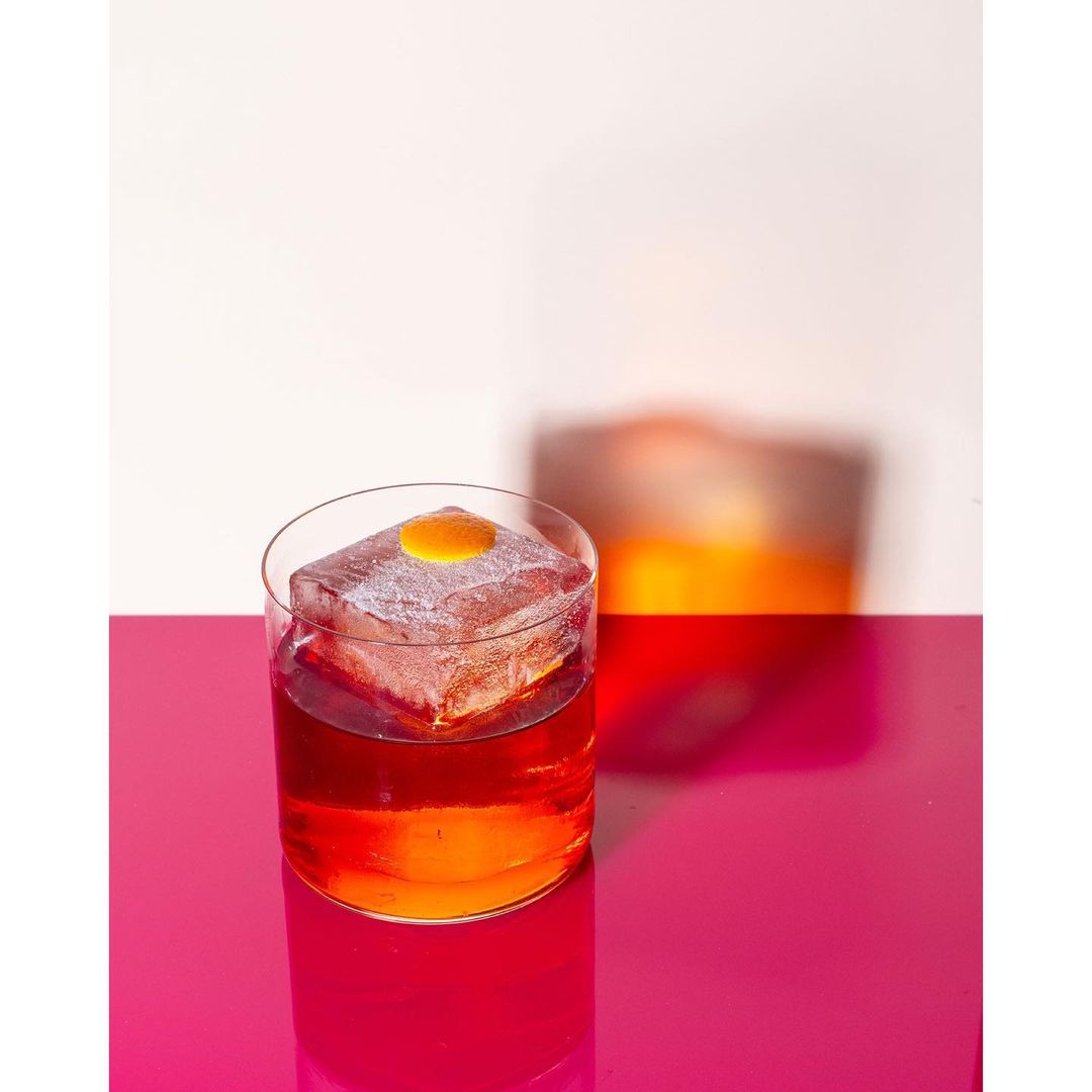 Cocktail in a rocks glass with large ice cube and orange zest garnish, resting on a shiny magenta surface, with a second glass in the background.