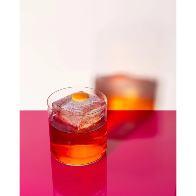 Cocktail in a rocks glass with large ice cube and orange zest garnish, resting on a shiny magenta surface, with a second glass in the background.