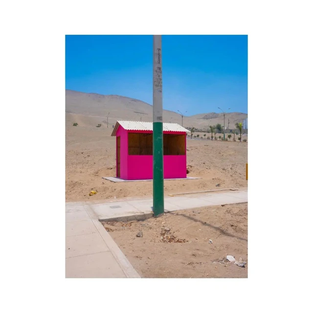 Hot pink roadside shack contrasts with a beige desert landscape under a clear blue sky, with a light gray concrete sidewalk.