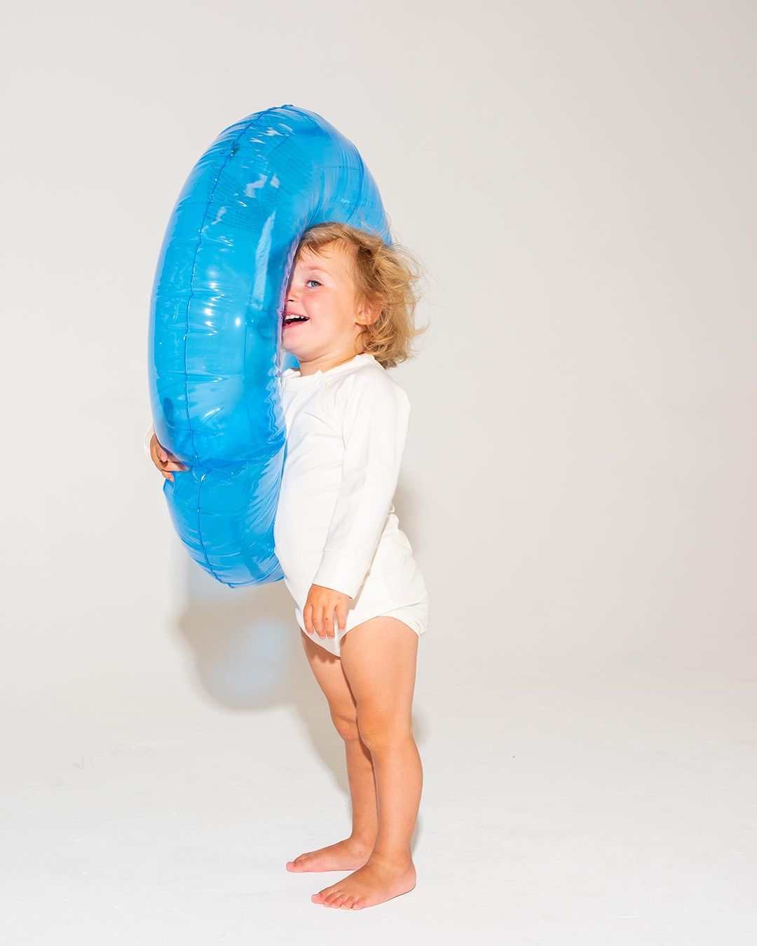 A child stands next to a large, shiny, blue, donut-shaped balloon against a plain white backdrop in a bright, eye-level shot.