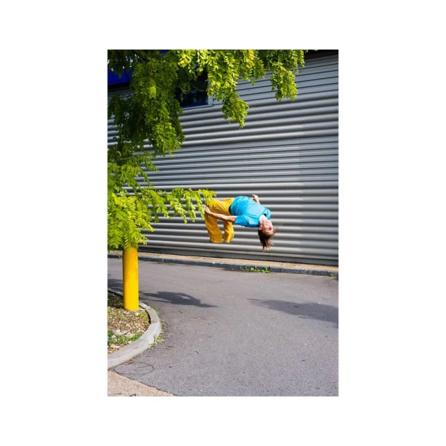 Adult performs gymnastic backbend near green tree, wearing blue t-shirt and yellow pants, on gray concrete with a corrugated metal wall.
