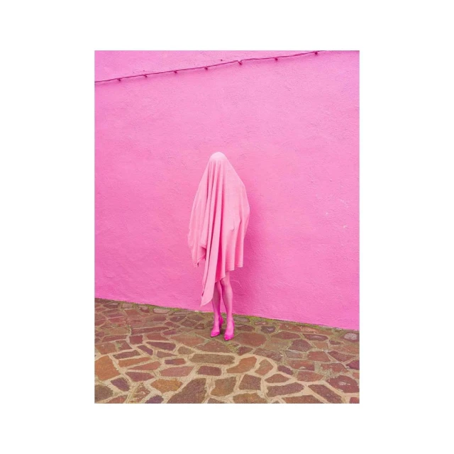 Person in pink draped fabric standing against a pink wall on a cobblestone surface; monochromatic fashion.