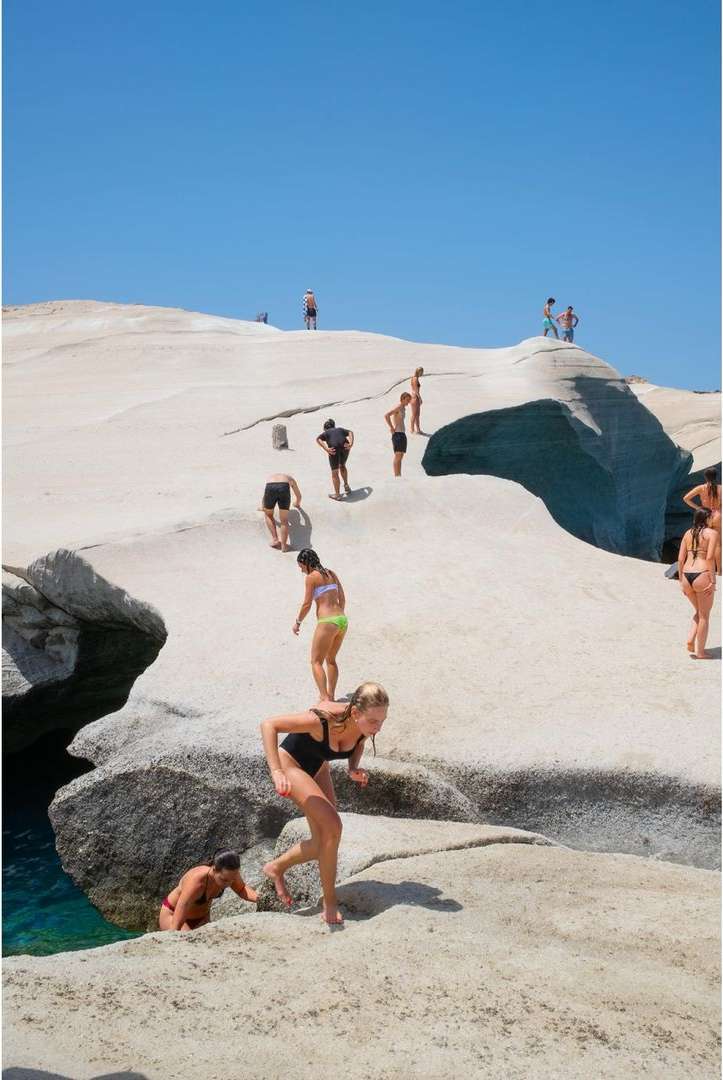 Multiple people on a rocky beach on a sunny day, with one woman in a black swimsuit in the foreground moving toward the water.