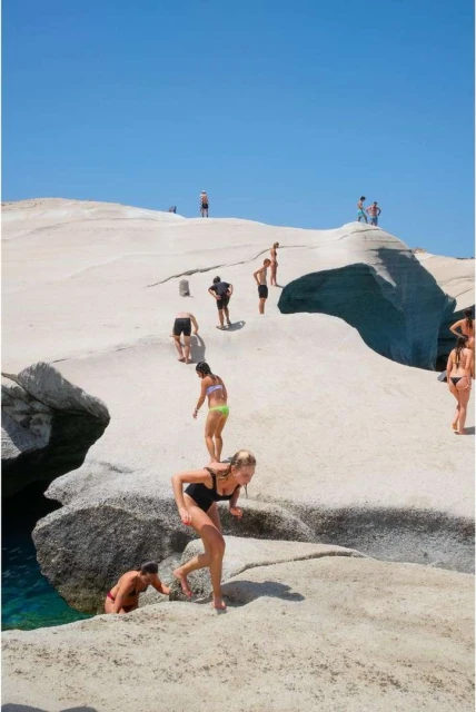 Multiple people on a rocky beach on a sunny day, with one woman in a black swimsuit in the foreground moving toward the water.