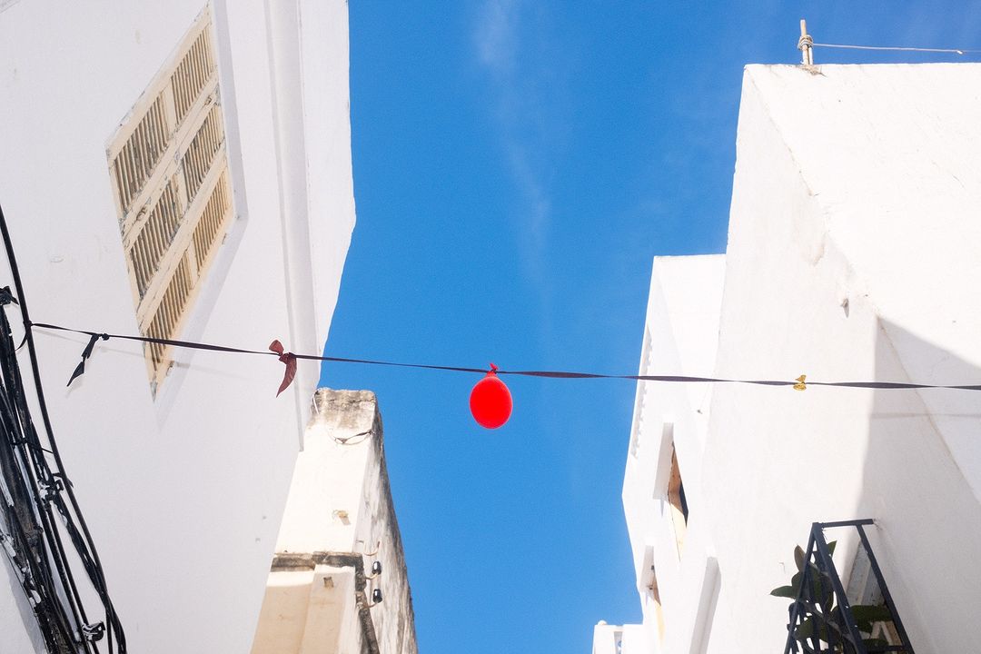 Low-angle view of a red balloon hanging between white buildings against a bright blue sky, creating a festive urban atmosphere.