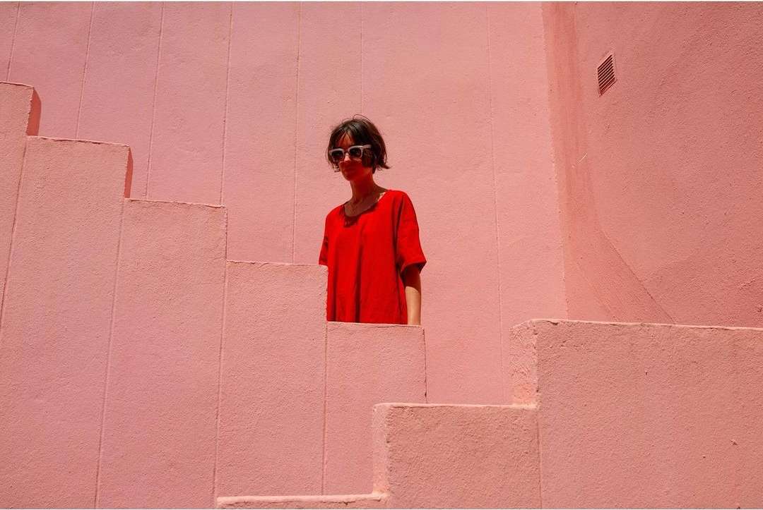 Young woman in sunglasses and a red shirt stands on a pink outdoor staircase bathed in light against a pink wall and background.