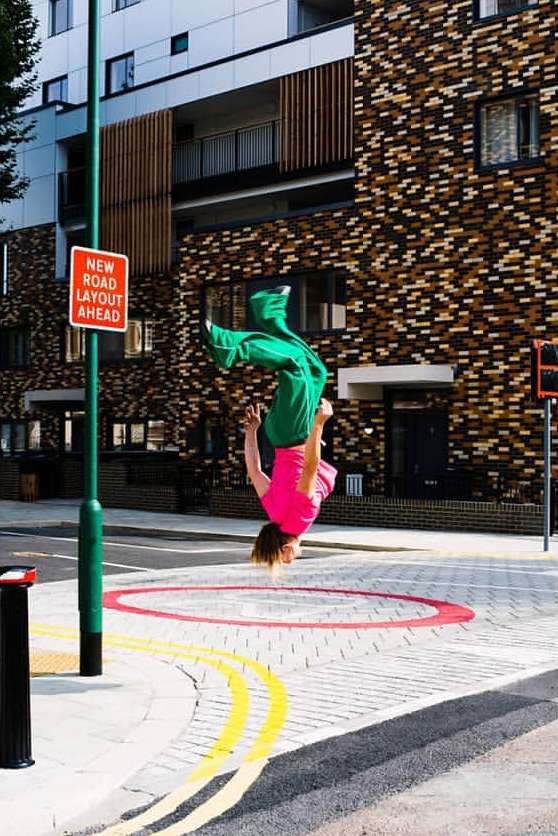 A person in green pants and a pink shirt performs a backflip near a building with a new road layout sign on a sunny day.