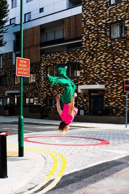 A person in green pants and a pink shirt performs a backflip near a building with a new road layout sign on a sunny day.