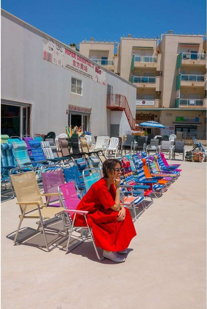 Woman in red dress sits on colorful folding chair outdoors amongst rows of chairs outside white building under bright sunny sky.
