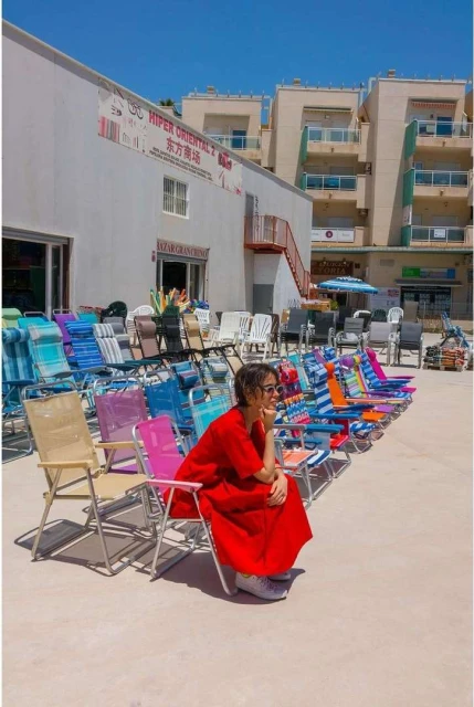 Woman in red dress sits on colorful folding chair outdoors amongst rows of chairs outside white building under bright sunny sky.