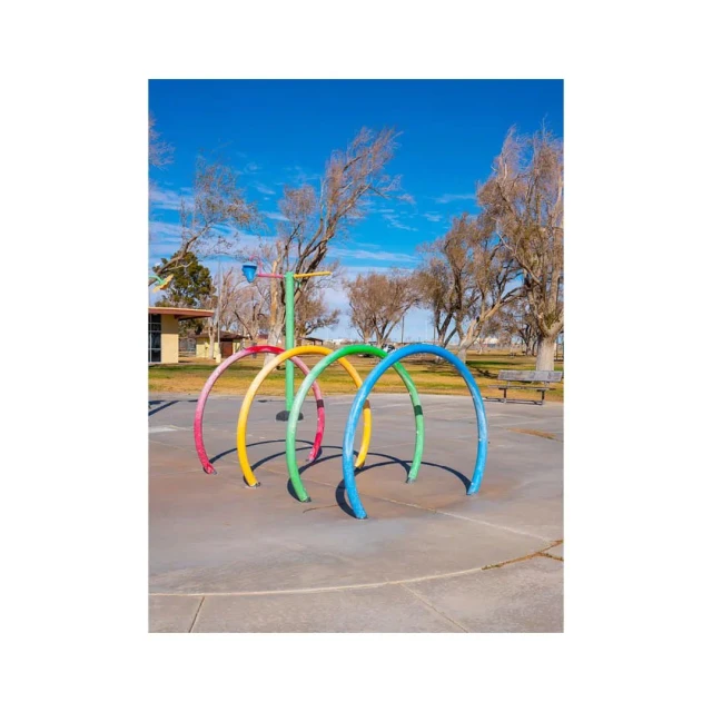 Colorful metal playground arches stand in a park under a blue sky near sparse trees and a building. Wide shot, daytime. 
