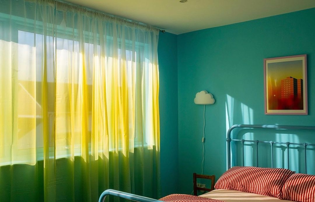 Sunlit bedroom interior with green and yellow curtains, a light blue metal bed frame, and turquoise walls featuring a framed print.