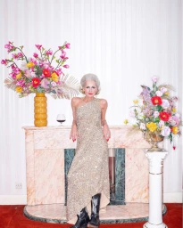 Older woman with silver hair wearing a sequined gown and cowboy boots standing before a marble mantelpiece with floral arrangements.
