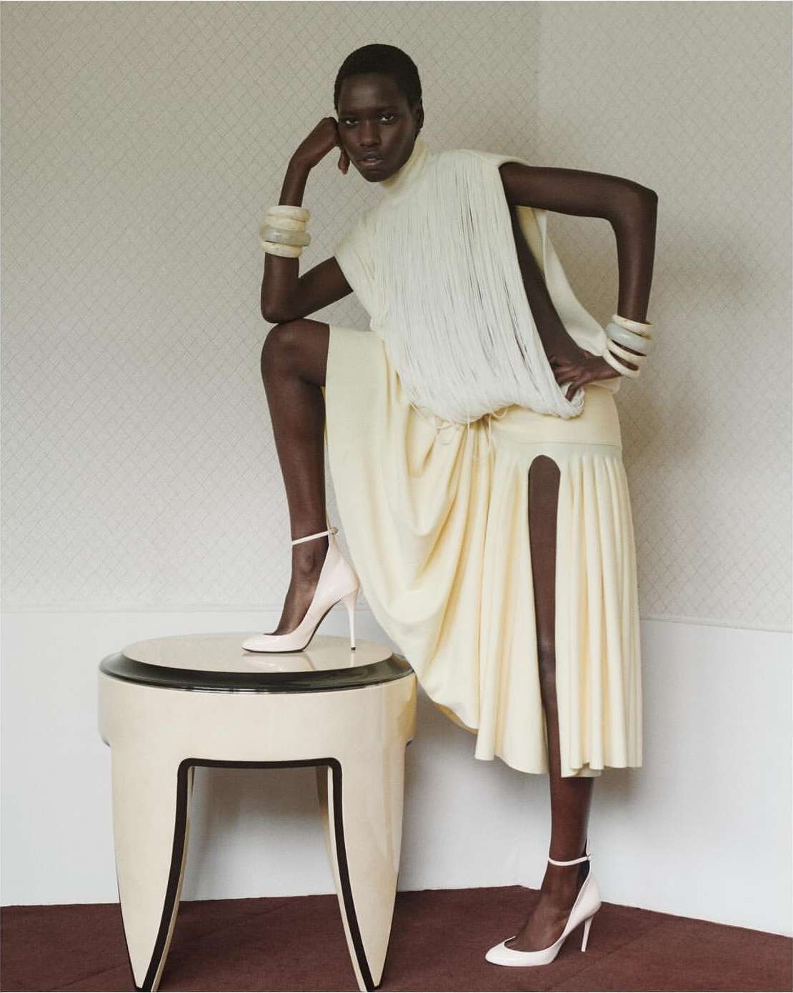 Elegant dark-skinned model in white dress and heels poses with leg on table, indoors, wearing chunky bracelets and high heels.