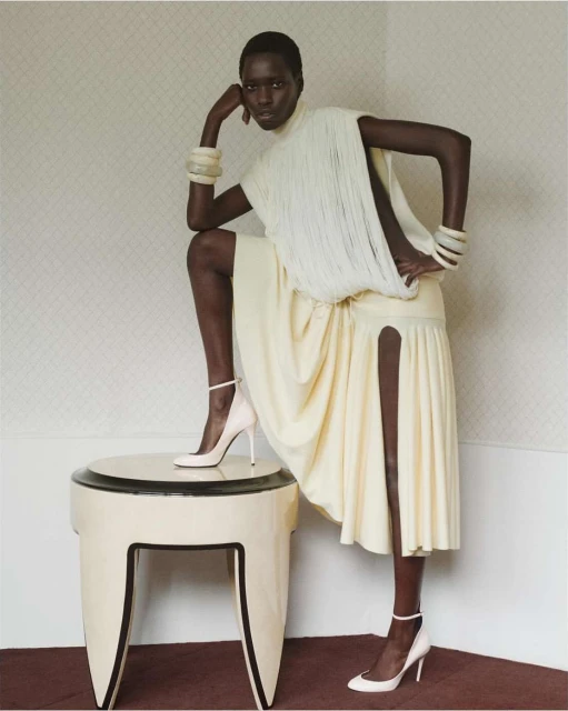 Elegant dark-skinned model in white dress and heels poses with leg on table, indoors, wearing chunky bracelets and high heels.