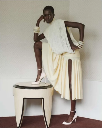 Elegant dark-skinned model in white dress and heels poses with leg on table, indoors, wearing chunky bracelets and high heels.