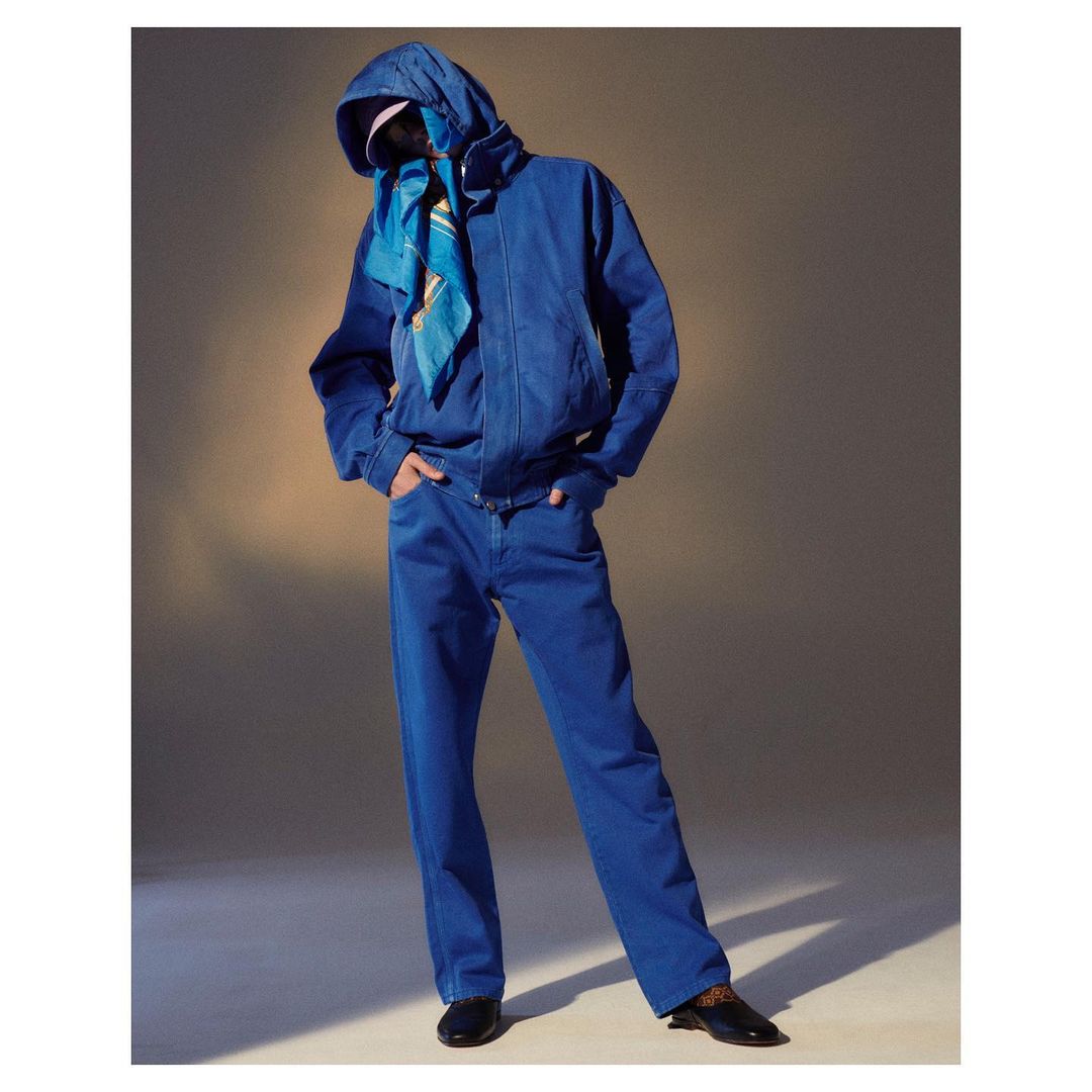 Person in blue jacket and jeans with a scarf stands in a studio with soft lighting. Fashion shot with covered face and modern style.