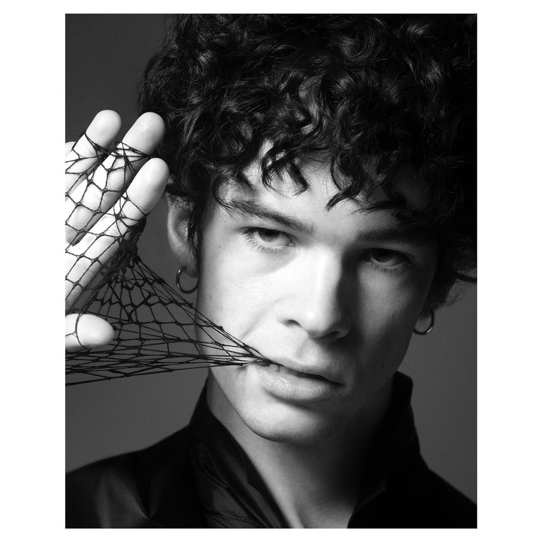 Black and white portrait of a young adult male with curly hair and an intense expression holding a black net glove near his mouth.