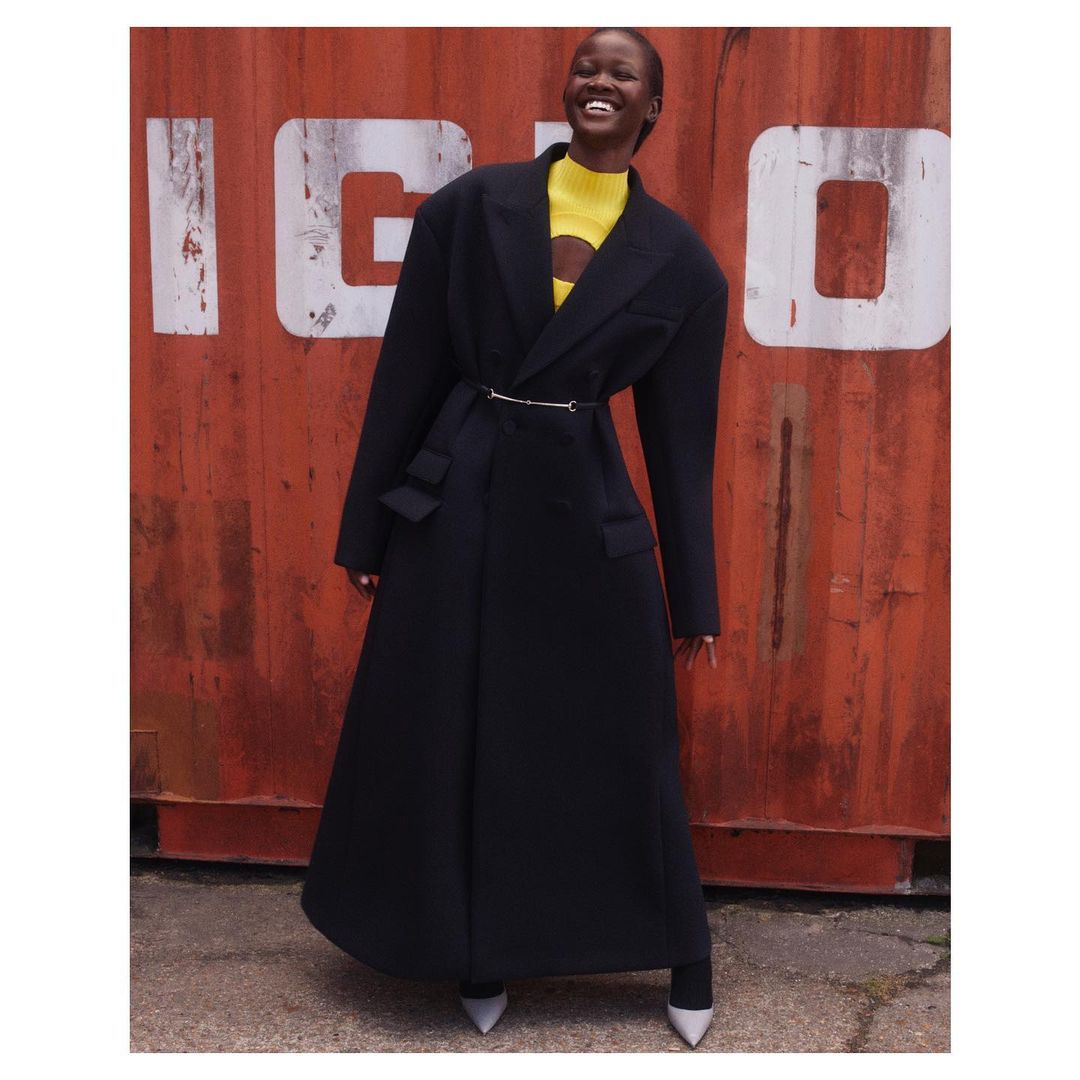 Smiling Black woman in a full-length black coat with a yellow top, posing outdoors in front of a rusty red corrugated metal wall.