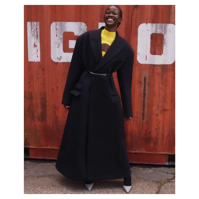 Smiling Black woman in a full-length black coat with a yellow top, posing outdoors in front of a rusty red corrugated metal wall.