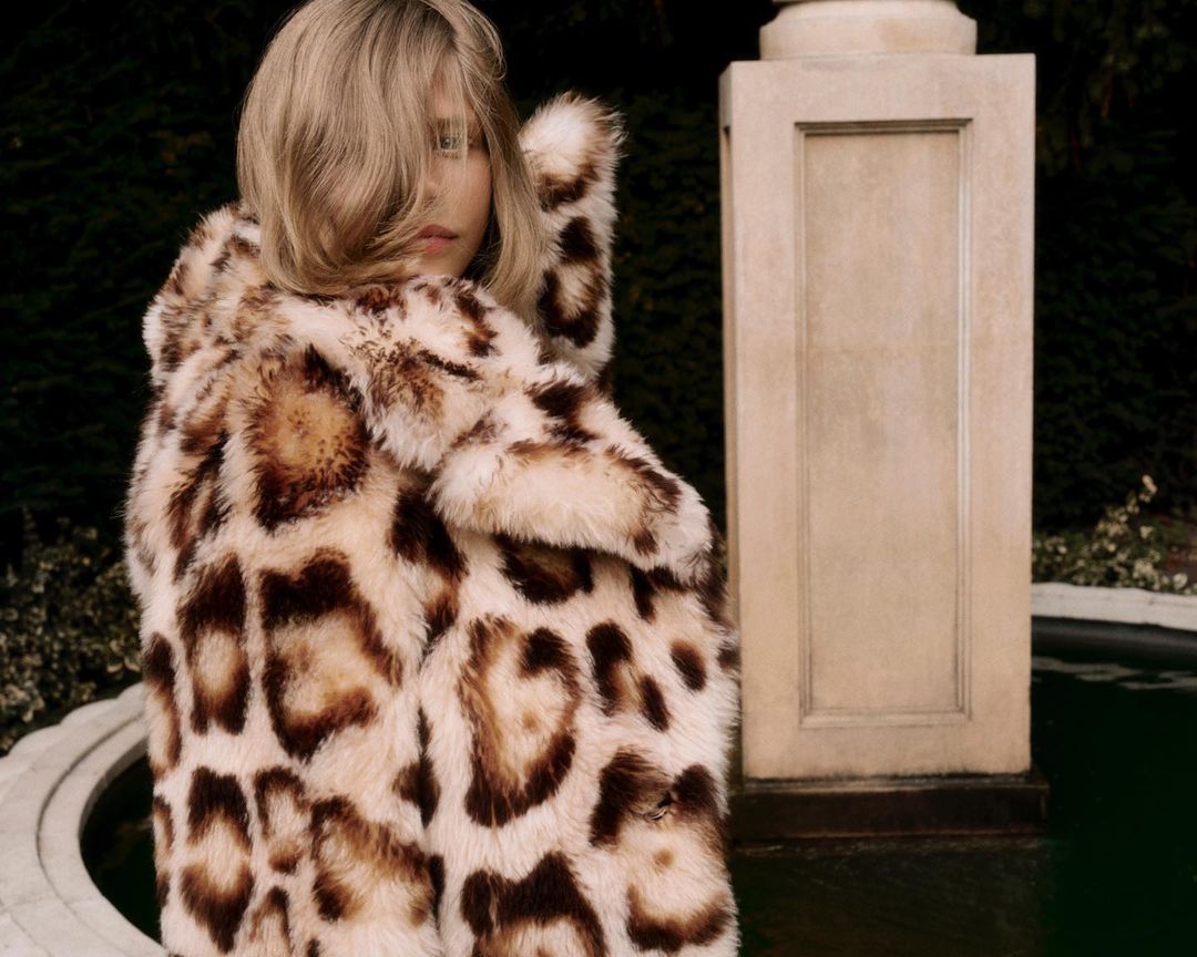 Adult female with blonde hair wearing a hooded faux fur coat with an animal print design standing outdoors near a stone column.