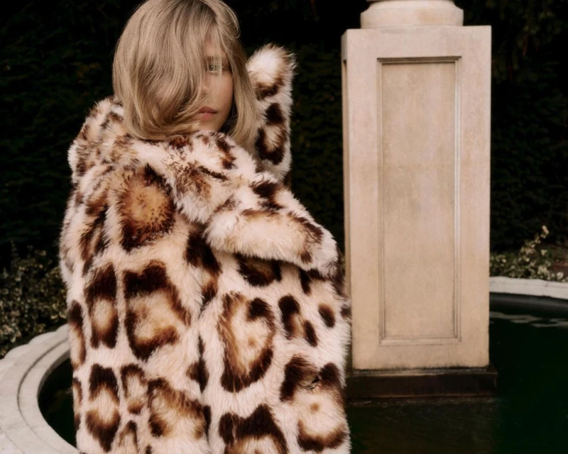 Adult female with blonde hair wearing a hooded faux fur coat with an animal print design standing outdoors near a stone column.