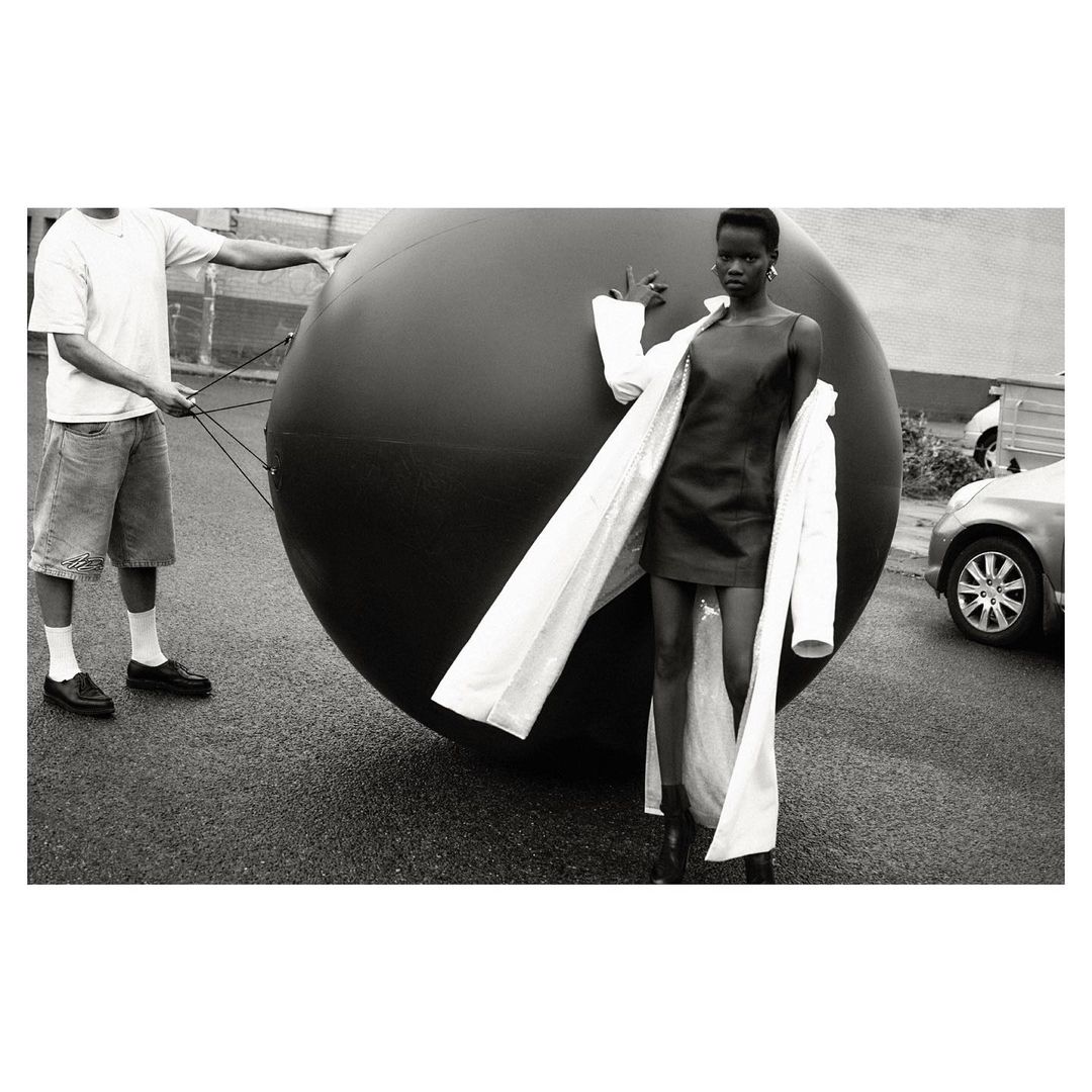 Black and white photograph of a Black woman posing in a black dress and white coat, standing in a street, next to a large black ball.