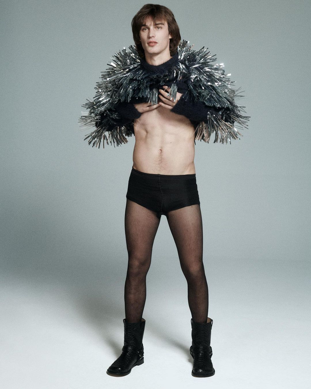 Man poses in black briefs, fishnet stockings, and platform boots, wearing a tinsel-covered cape in a light grey photo studio.
