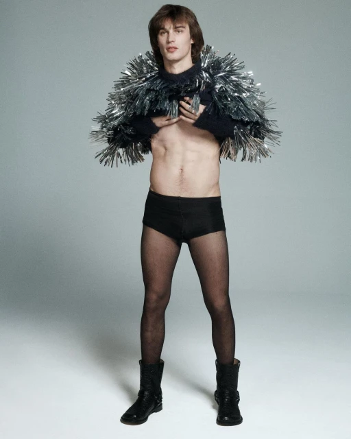 Man poses in black briefs, fishnet stockings, and platform boots, wearing a tinsel-covered cape in a light grey photo studio.