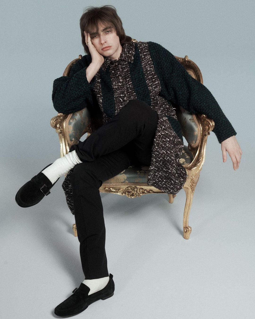 Young man with dark hair seated in an ornate armchair wearing a patterned cardigan and black trousers by Will Scarborough