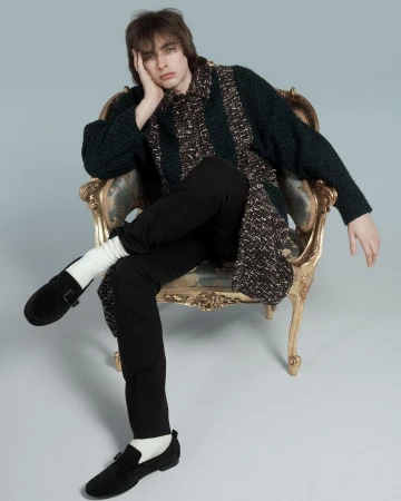 Young man with dark hair seated in an ornate armchair wearing a patterned cardigan and black trousers by Will Scarborough