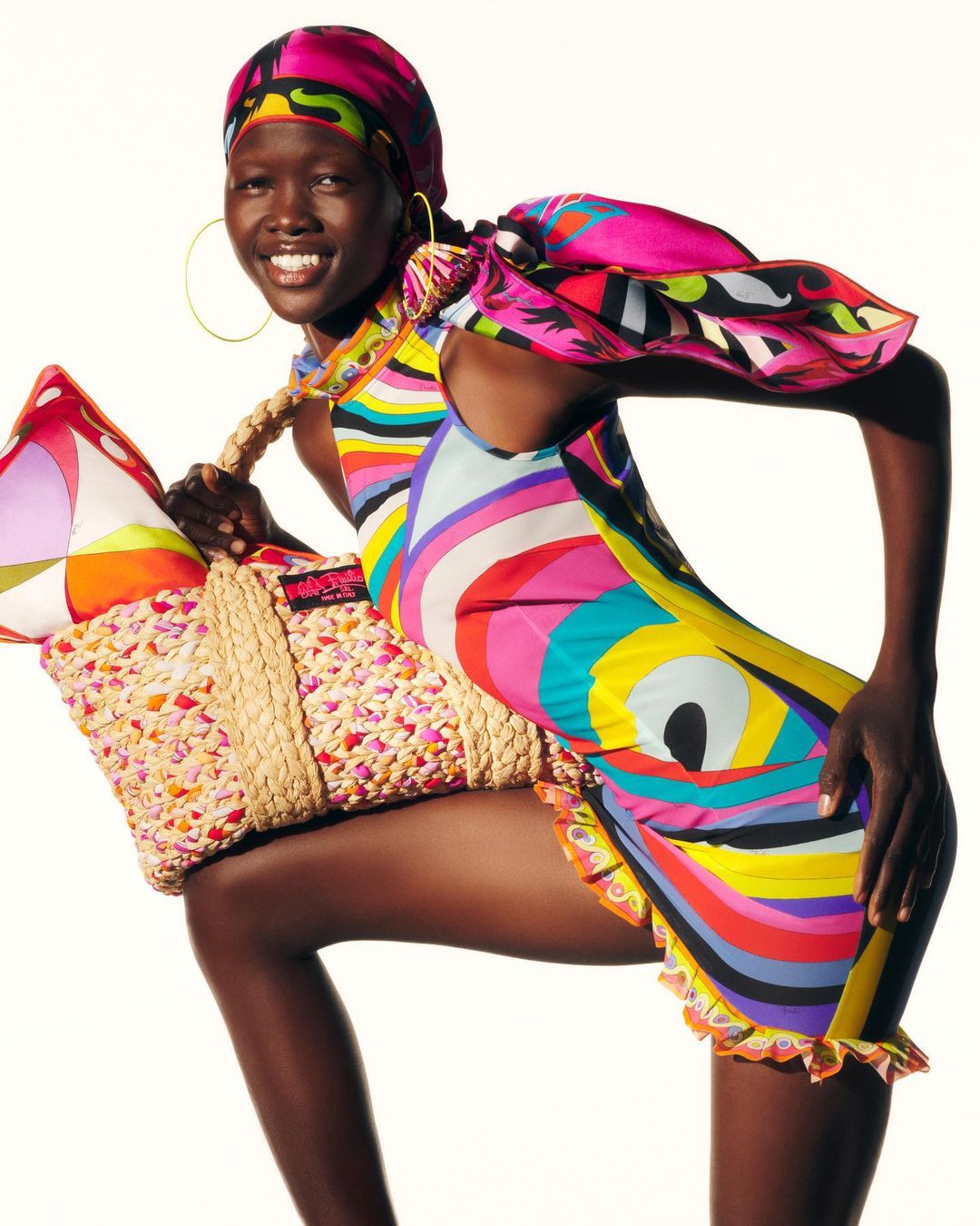 Fashion shot featuring a smiling woman wearing a bright, patterned mini dress and headscarf, carrying a woven tote bag on a white background.