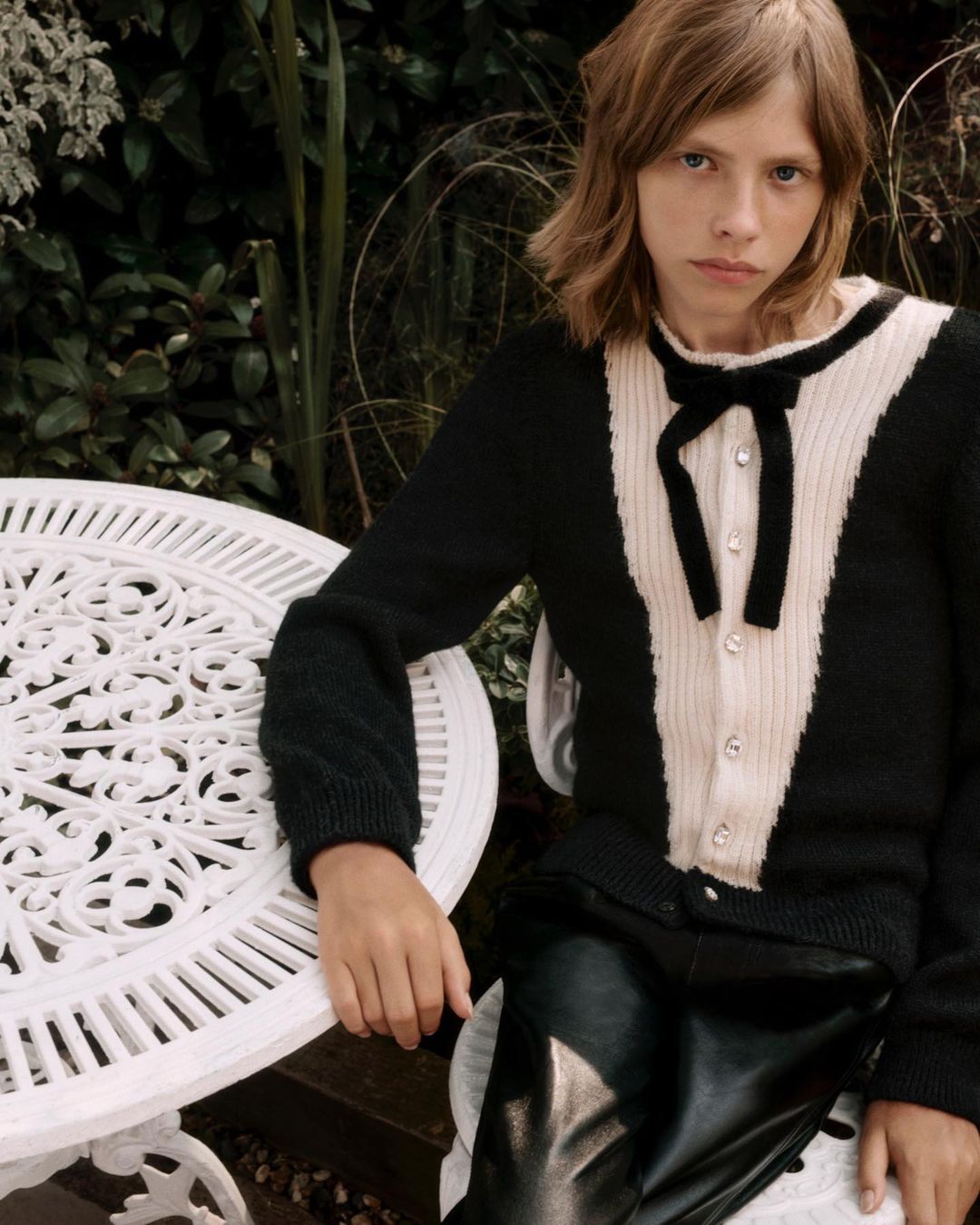 Model with long reddish-brown hair wearing a black and cream cardigan with a bow, seated outdoors by a white table.