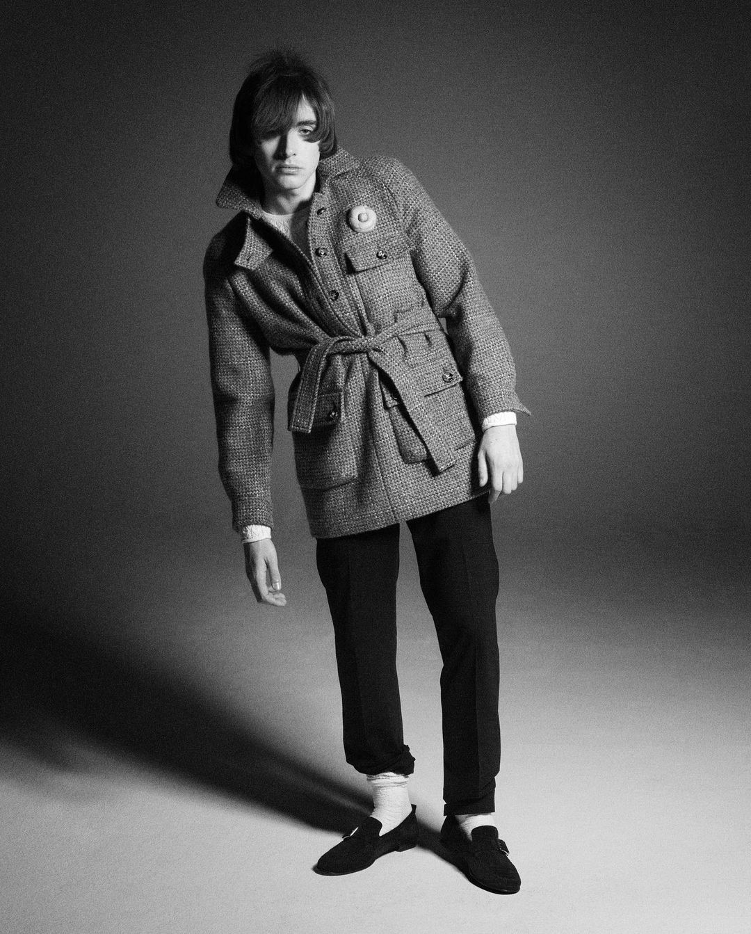 Black and white portrait of a stylish young adult male in a tweed belted jacket, trousers, and loafers, posing in a studio setting.