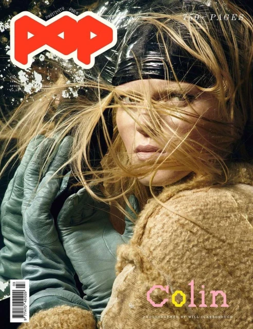 Magazine cover featuring a blonde woman with hair partially covering her face, wearing a black cap, tan coat, and blue gloves.