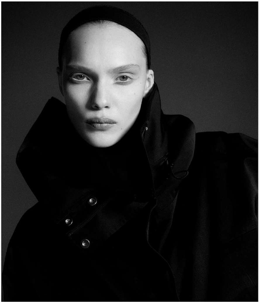 Monochrome portrait of a fair-skinned woman with a black headband and high-collared coat against a dark grey background.