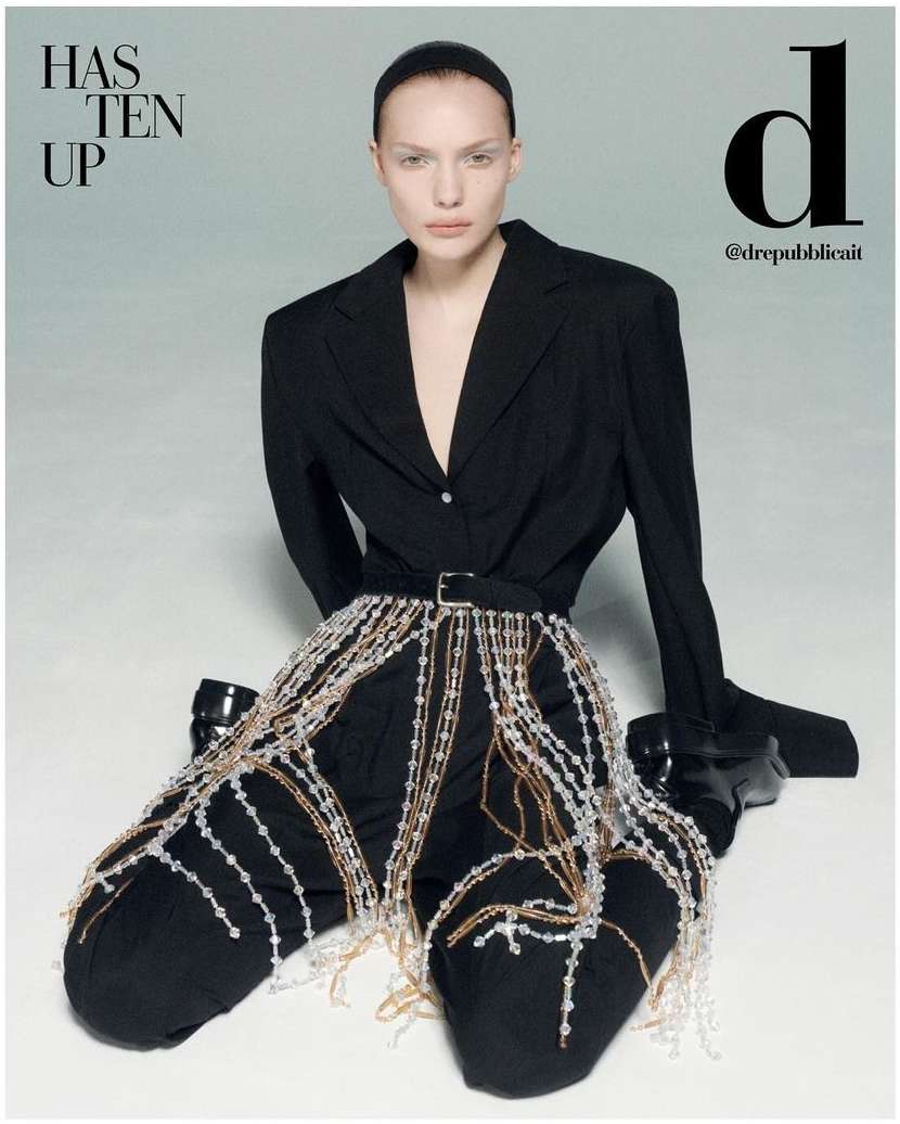 A fashion editorial shot features an adult female model in a black suit with beaded embellishments against a light grey background.