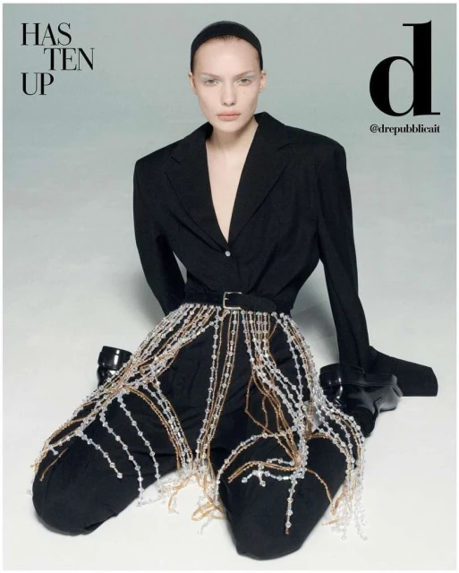 A fashion editorial shot features an adult female model in a black suit with beaded embellishments against a light grey background.