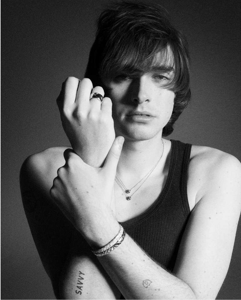 Portrait of a man in a black tank top holding his wrist showcasing rings and bracelets against a grey backdrop
