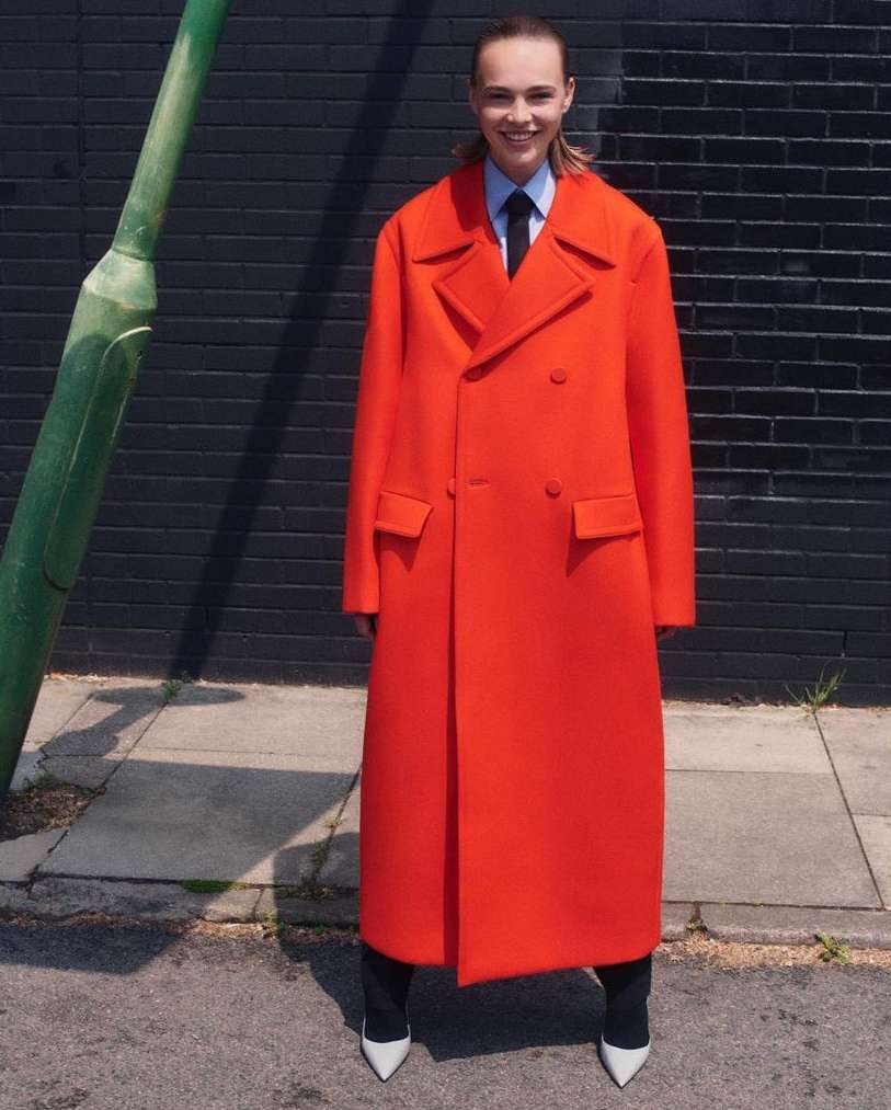 A woman with fair skin and brown hair smiles while wearing an orange overcoat, a blue dress shirt, and white high heels outdoors on a street.