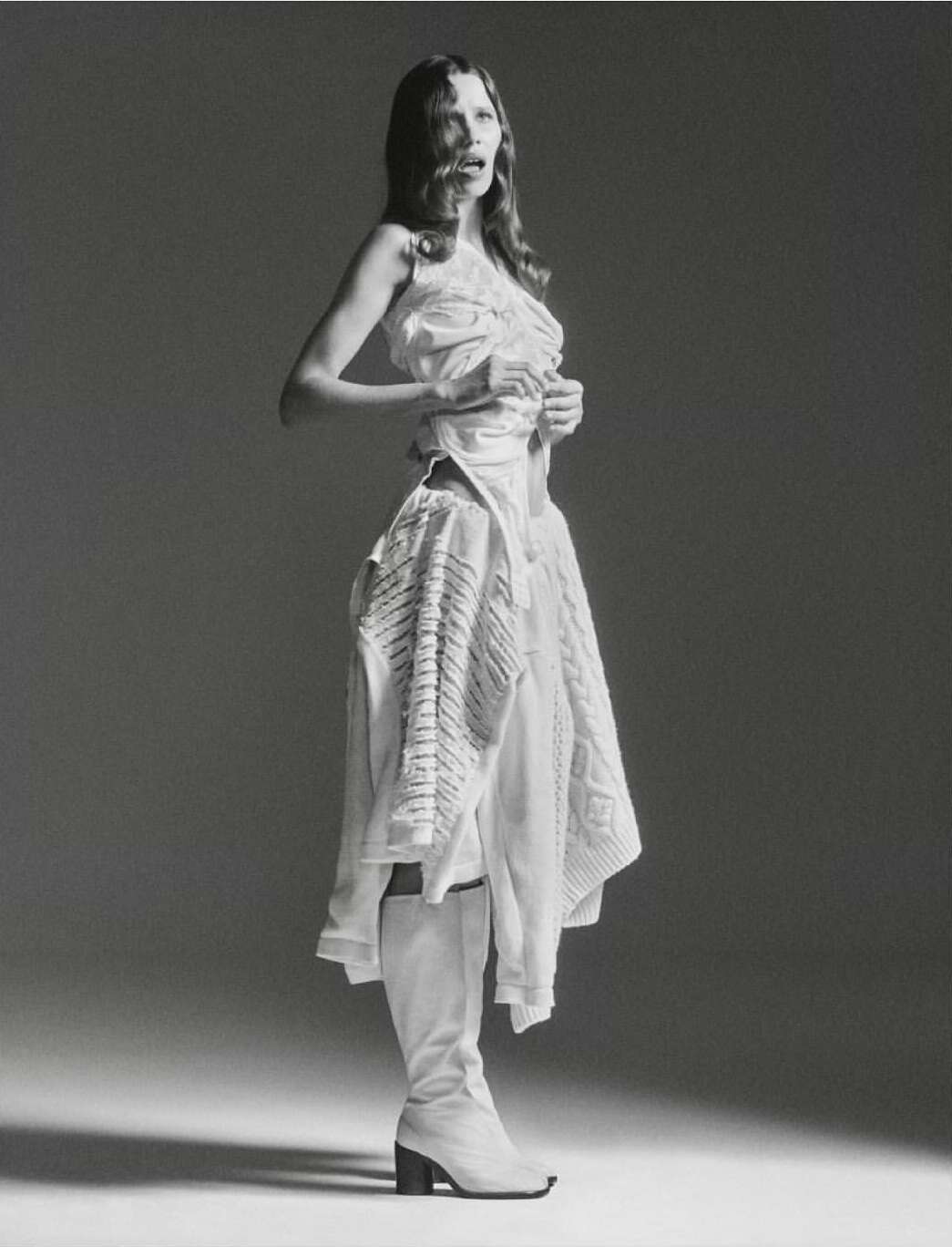 Fashion shot of a woman in a white dress and boots, posing in a photo studio with a neutral backdrop and soft lighting for editorial use.