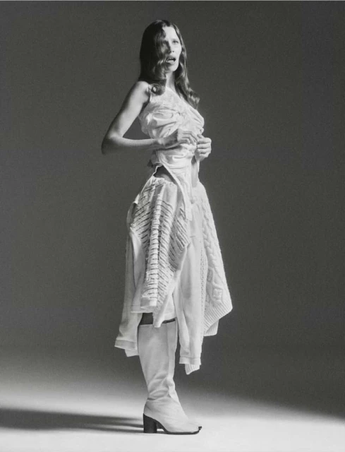 Fashion shot of a woman in a white dress and boots, posing in a photo studio with a neutral backdrop and soft lighting for editorial use.