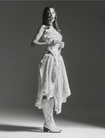 Fashion shot of a woman in a white dress and boots, posing in a photo studio with a neutral backdrop and soft lighting for editorial use.