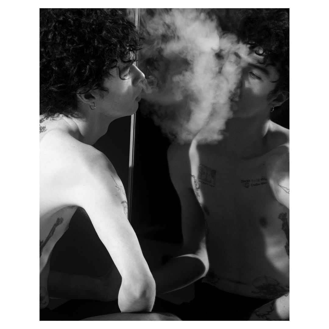 Monochrome portrait of a shirtless person exhaling smoke in front of a mirror, capturing both the individual and their reflected image.